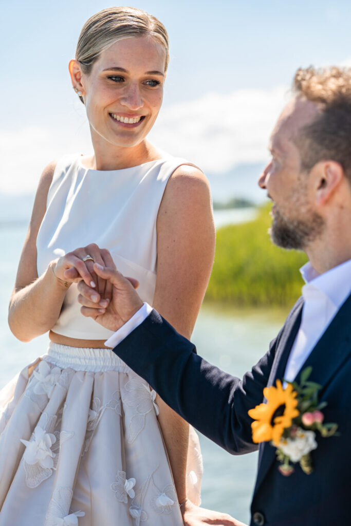 Brautpaar hält Händchen vor Chiemsee-Kulisse, Braut in modernem zweiteiligem Outfit